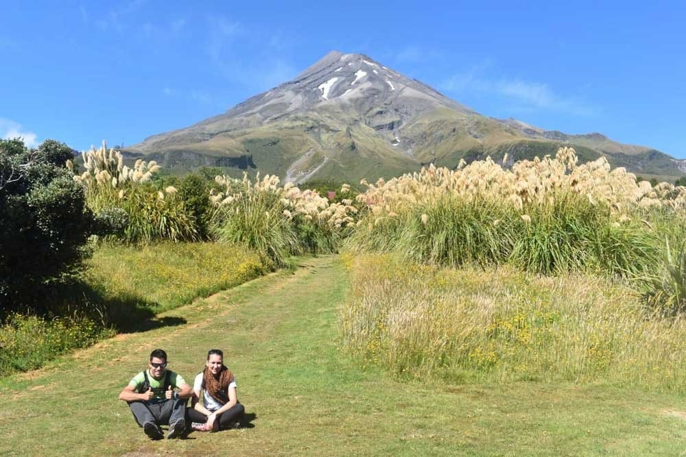 Viajar a Nueva Zelanda chico morena y chica sentados en el césped mirando a cámara y detrás una montaña